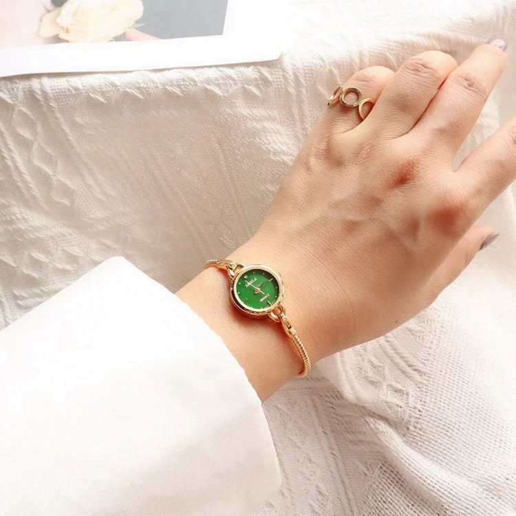 Gold bracelet watch with green face on wrist, paired with gold ring, white sleeve visible