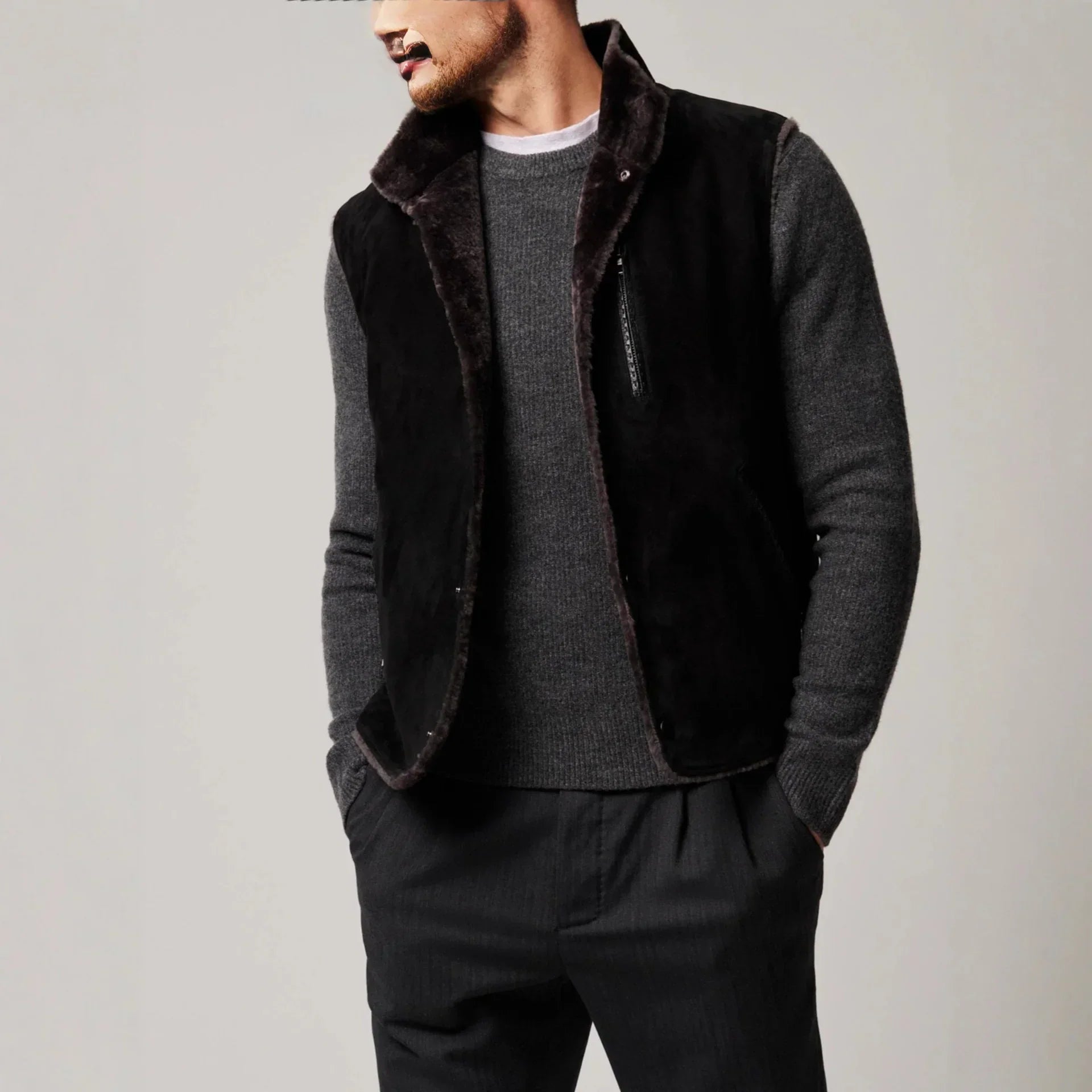 Man wearing a black faux fur vest over a grey knit sweater and dark tailored pants