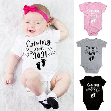 Baby wearing white cotton onesie with 'Coming Soon 2021' print, available in pink, grey, and black.