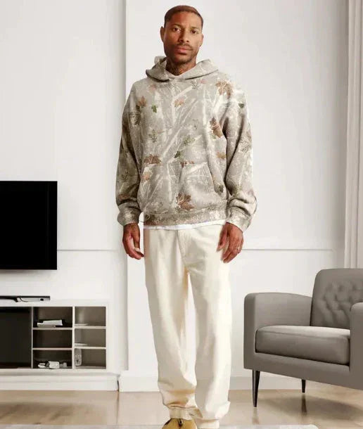 Man wearing a relaxed fit, nature-print hoodie and cream straight-leg pants in a modern living room.