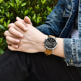 Man wearing blue denim jacket, black pants, and a brown leather strap watch outdoors