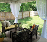 Outdoor patio set with dark wood chairs, yellow and beige cushions, white sheer curtains, and decorative pillows.