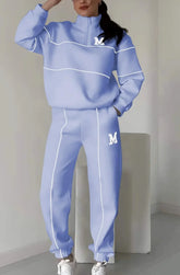 Light blue streetwear tracksuit with white piping, relaxed fit, and zip neck, worn indoors.