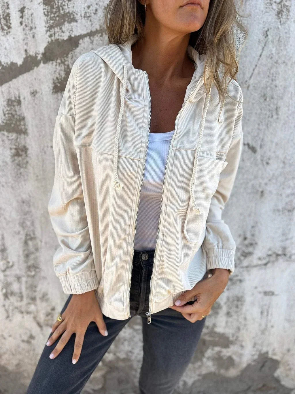 Woman wearing a cream corduroy zip-up hooded jacket, white ribbed tank top, and black jeans