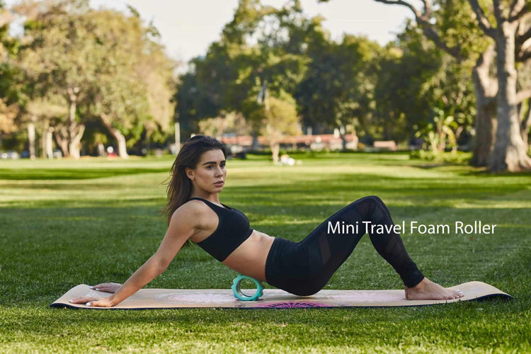 Woman in black fitted workout leggings and sports bra using a mini foam roller outdoors on a yoga mat